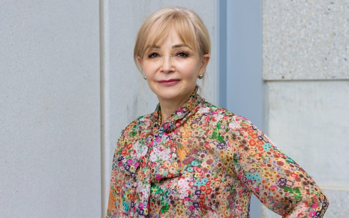 Portrait photograph of a woman with blonde hair, wearing a colourful floral shirt and with one hand on her hip. 