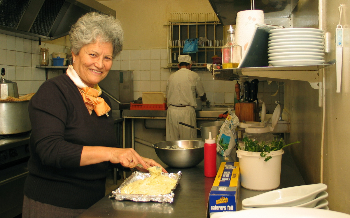 Woman with short grey hair smiling at the camera as she spreads food in a foil-lined tray in the kitchen of a restaurant