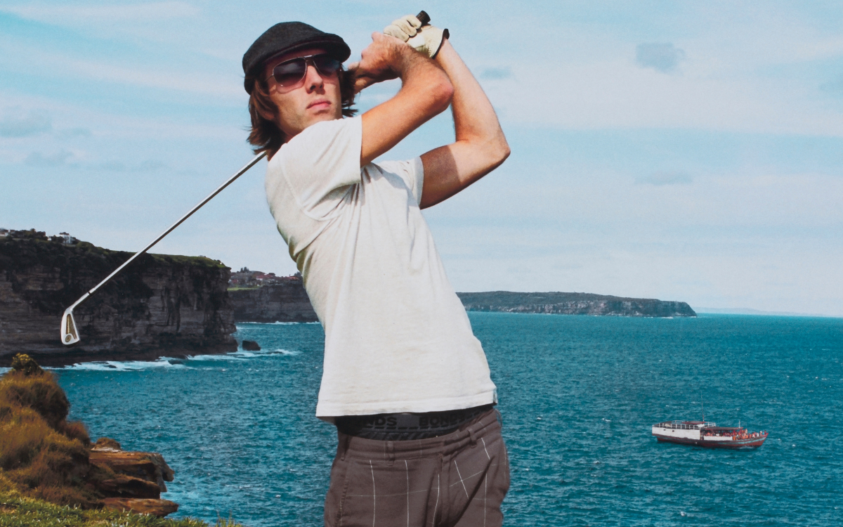 Man in a polo shirt, shorts, a hat and sunglasses playing golf near a cliff over the ocean