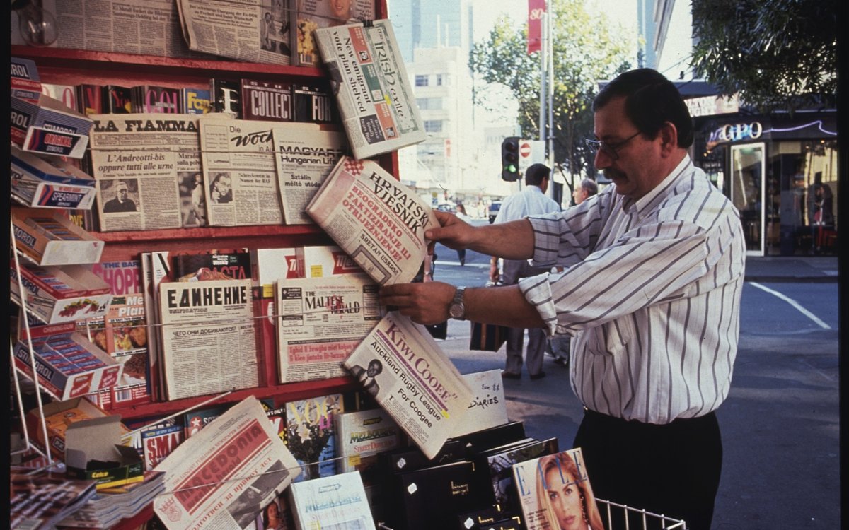 Man in striped button up shirt tucked into dark pants looking at newspapers in various languages at a kiosk on the street