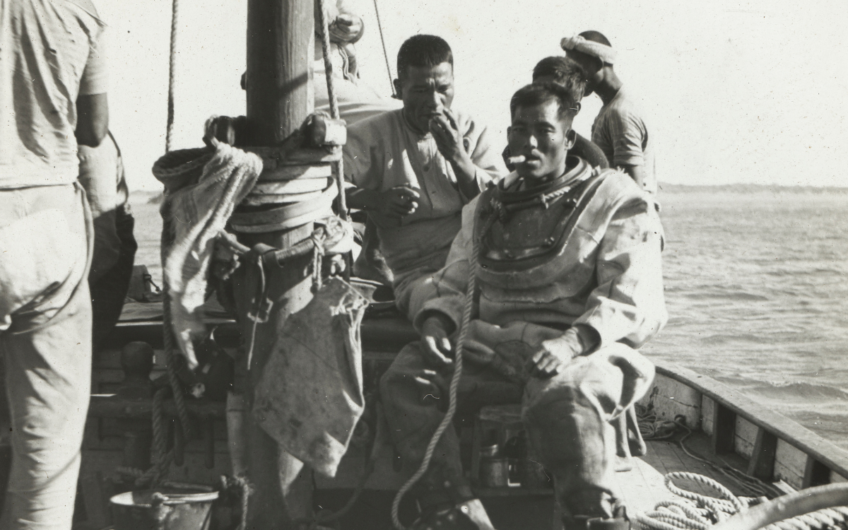 Black and white photo of Japanese men on a pearl diving boat, one dressed in diving attire but with the helmet off