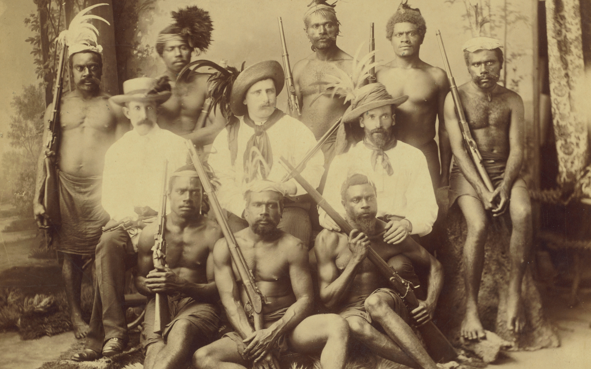 Group portrait of eight Pacific Islander men holding large guns and three British 'recruiters'