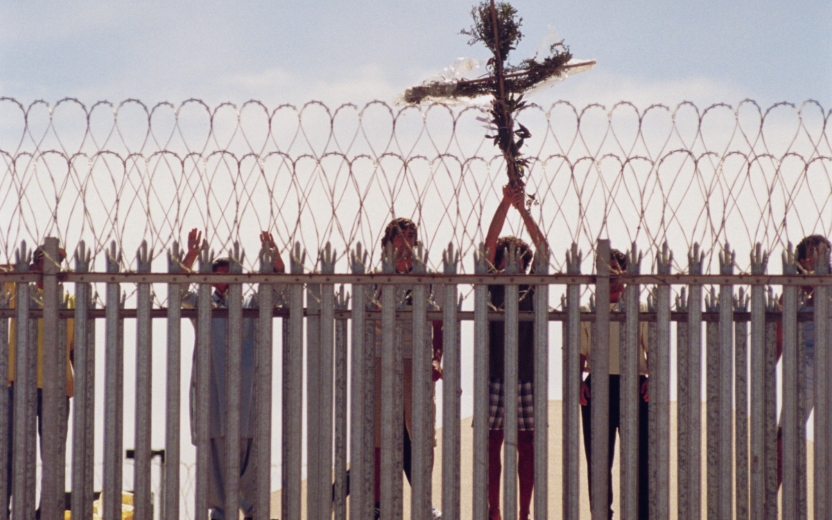 People standing behind a tall fence topped with barbed wire, one of whom is holding up a cross