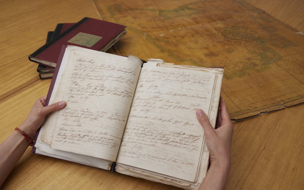 Hand holding an old notebook with handwritten text. In the background is a stack of books and an old map.