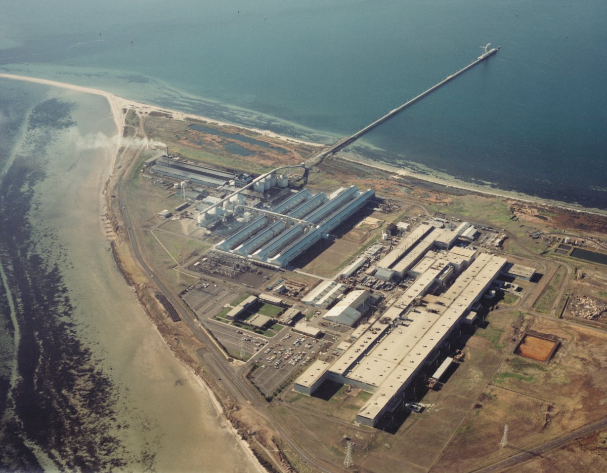 An aerial shot of a huge factory on a point surrounded by ocean. The factory is made up of several large sheds and warehouses. A long pier runs out into deep water.