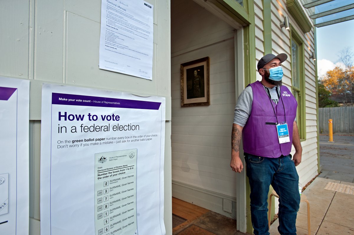 A photograph of a man wearing a purple vest that says "AEC STAFF" and a blue medical facemask. He is standing in front of a building one which posters explaining how to vote in a federal election are hanging.