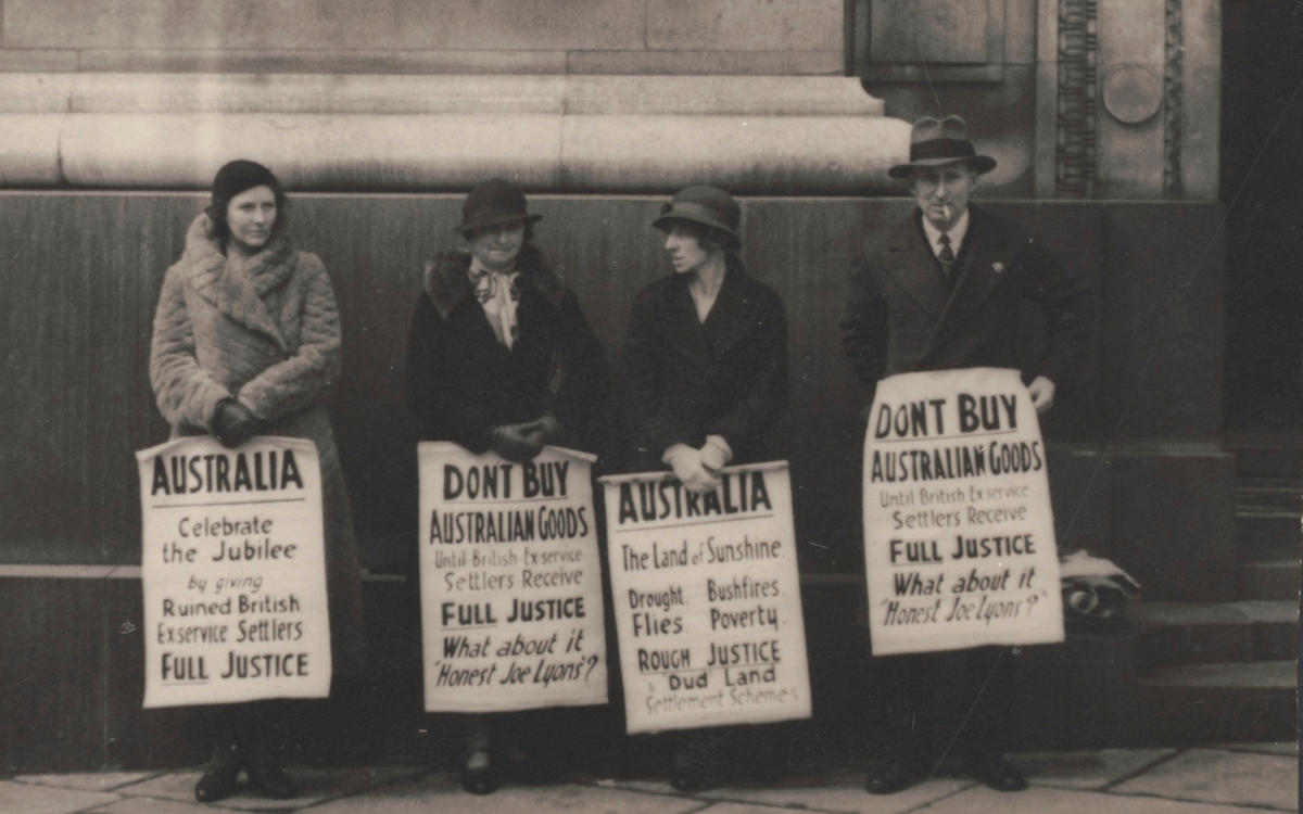 Black and white photo of three women and one man in winter clothes standing outside a building with signs protesting the treatment of British soldier settlers in the Australian War Service Land Settlement Scheme