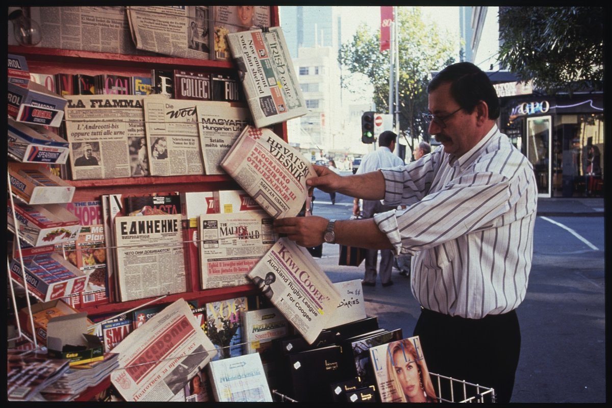Man in striped button up shirt tucked into dark pants looking at newspapers in various languages at a kiosk on the street