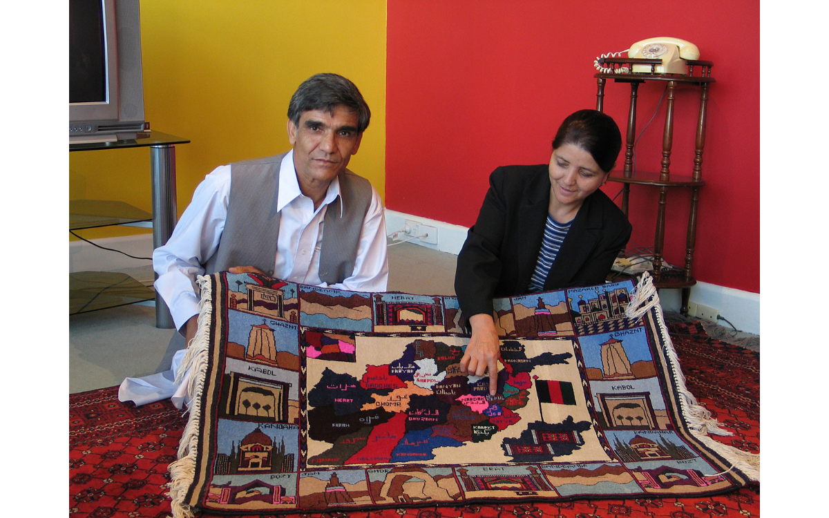 Man and woman of Middle Eastern heritage sitting on the floor, holding and looking at a rug with a map of Afghanistan and it's landmarks