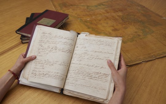 Hand holding an old notebook with handwritten text. In the background is a stack of books and an old map.