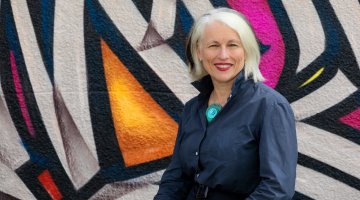 A portrait image of Genevieve Jacobs AM standing in front of a colourful wall