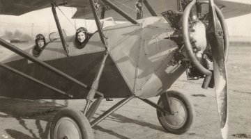A black and white image of two people sitting in a 1920s style aeroplane while grounded.