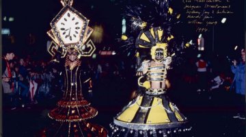 Two people in a night time parade dressed up in large, intricate, colourful costumes.