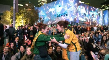 Two woman in green and gold clothing embracing excitedly in front of a crowd.