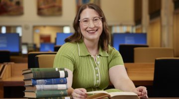 Zoe Smith, an NLA Scholar, sitting at a table in the Main Reading Room with a pile of books and smiling