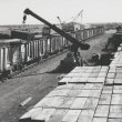 A sepia image of a crane loading pallets of sleepers from a stockpile onto a train for the railhead of the new 265 mile railway from Mount Newman and Mount Whaleback to Port Hedland, Western Australia in August 1968.