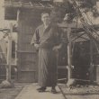 A sepia photograph of a man in traditional Japanese dress standing outside in front of a wooden dwelling structure.