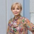 Portrait photograph of a woman with blonde hair, wearing a colourful floral shirt and with one hand on her hip. 