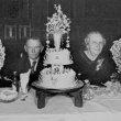 Husband and wife sitting at a table with a large cake and vases of flowers in front of them