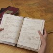 Hand holding an old notebook with handwritten text. In the background is a stack of books and an old map.