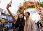 Young man and woman smiling and posing for a selfie in front of a large flower arch with a neon sign reading 'I heart CABRA'