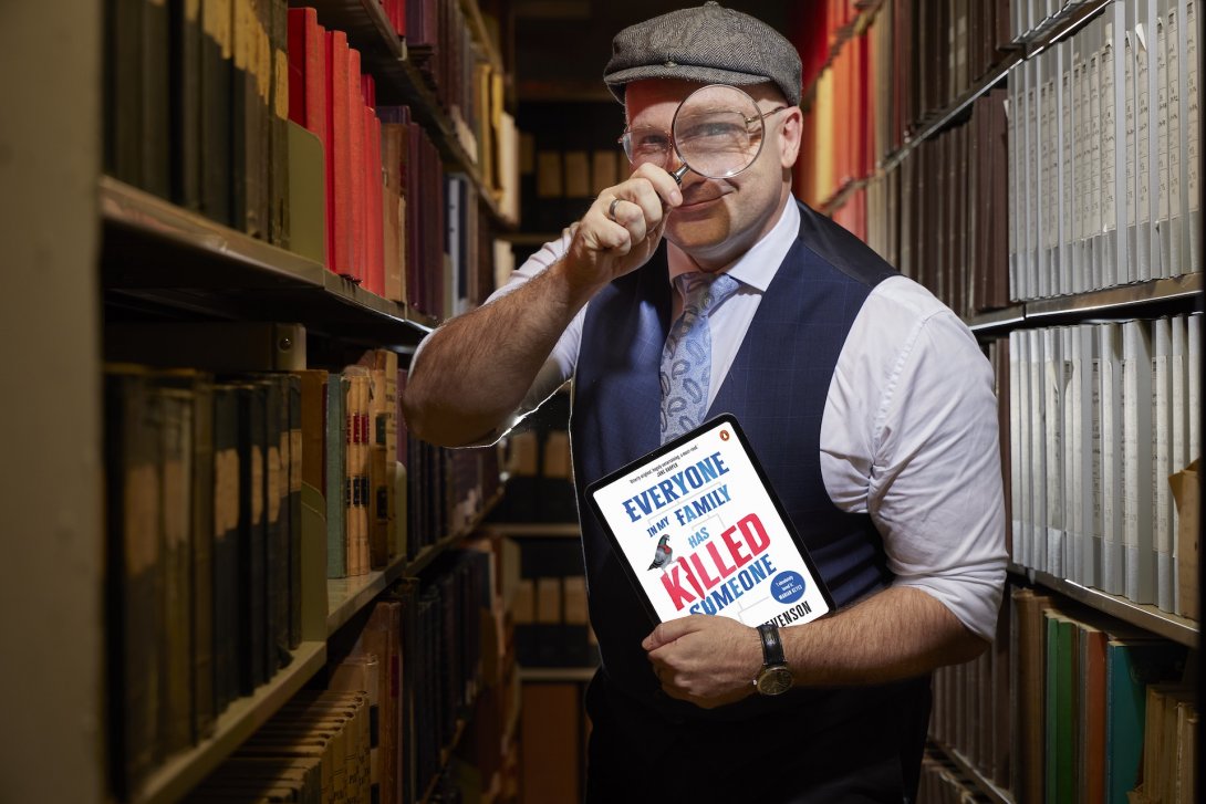 Man standing in the stacks of the Library wearing a button up shirt, tie, waistcoat, and newsboy cap holding a tablet displaying a book cover in his left hand and holding a magnifying glass up to his eye with his right hand. 