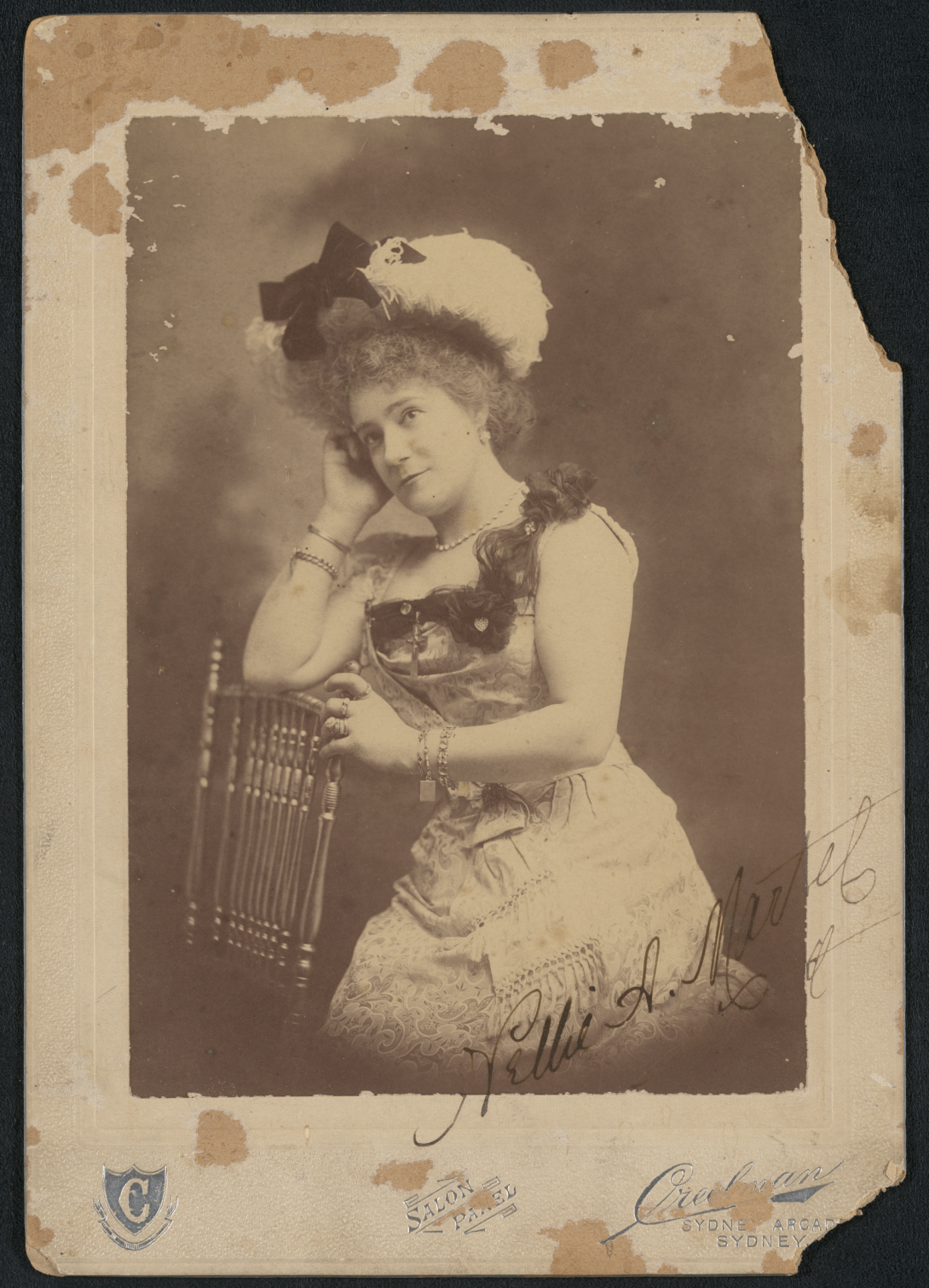 Old photograph of a woman with curly hair, around hat and a 1900s style dress leaning on a chair