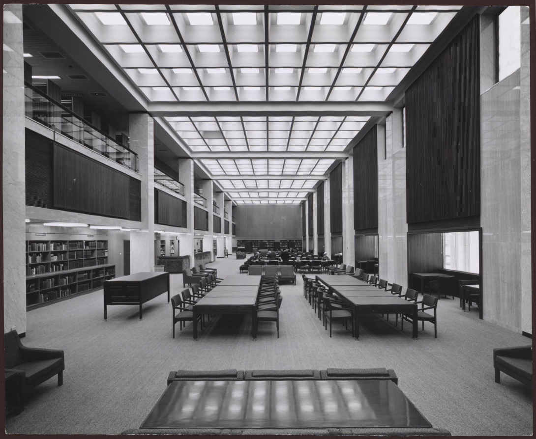 Black and white photo of a large, tall room with lights covering the ceiling, large tables with chair and bookshelves along the wall 