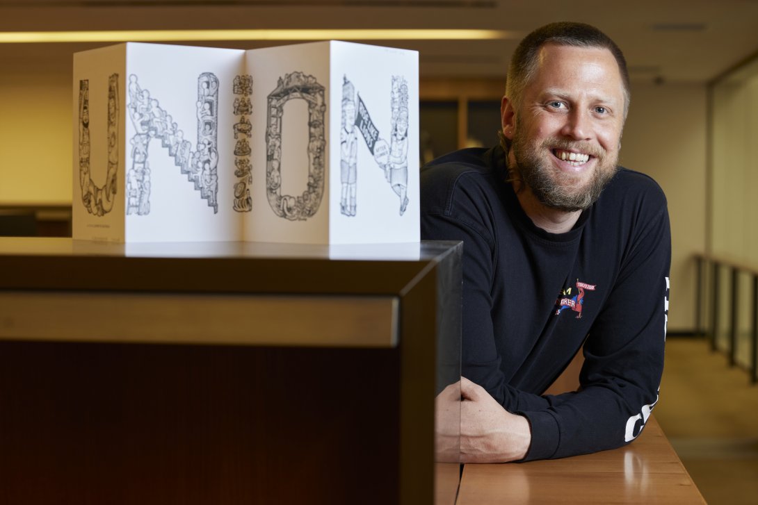 Smiling man leaning on a wooden surface behind a fold-out artwork reading 'UNION'