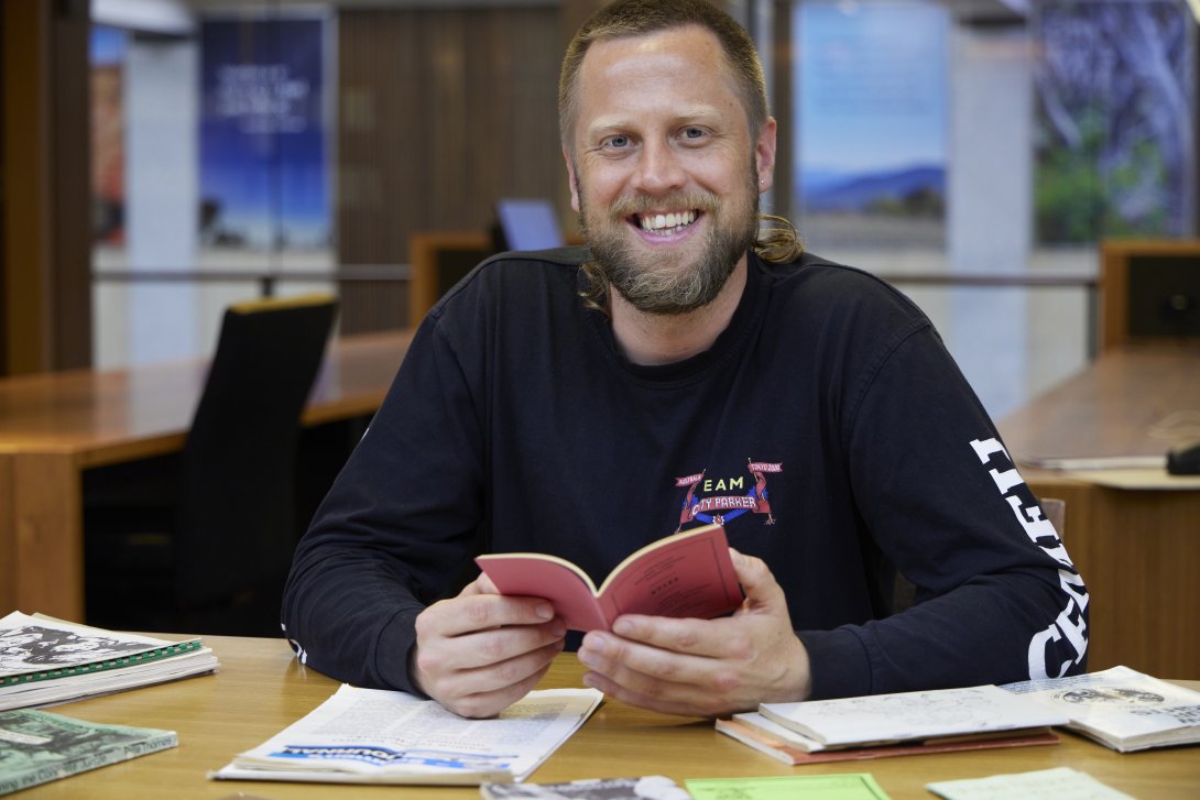 Man smiling as he holds a small red booklet and sits at a desk covered with books and papers
