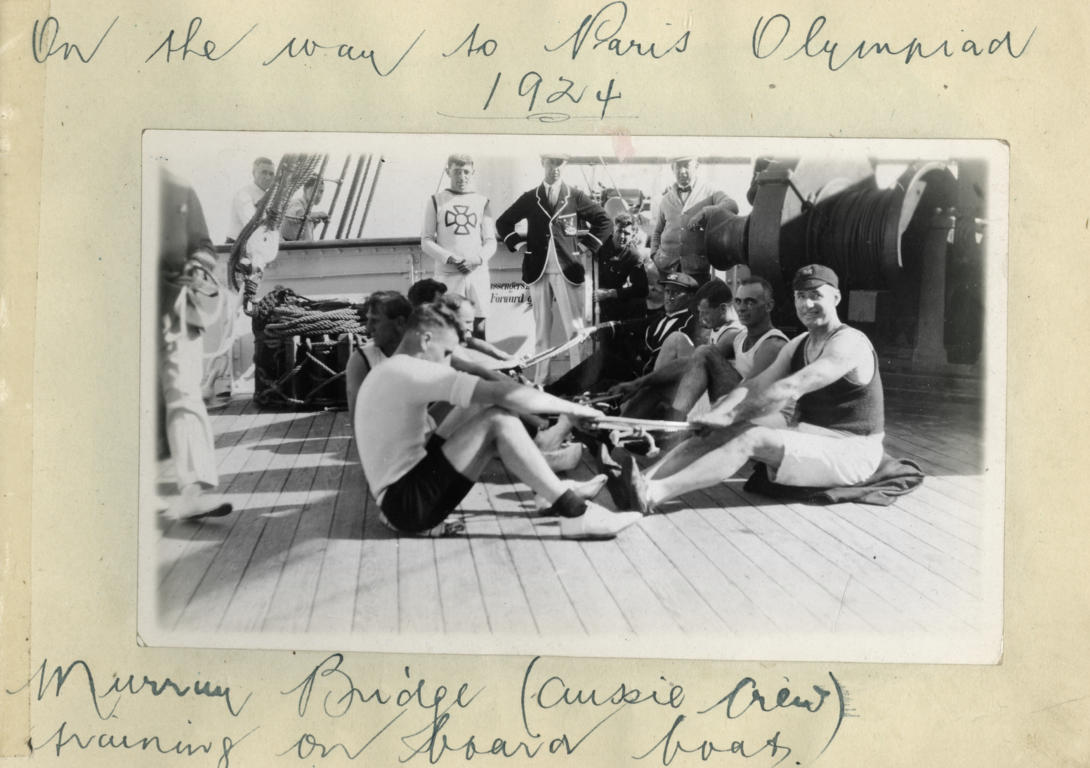 Black and white photo of men sitting and training on the deck of a ship stuck in a book with handwriting reading 'On the way to Paris Olympics 1924' on above and 'Murray Bridge (aussie crew) training on board boat' below