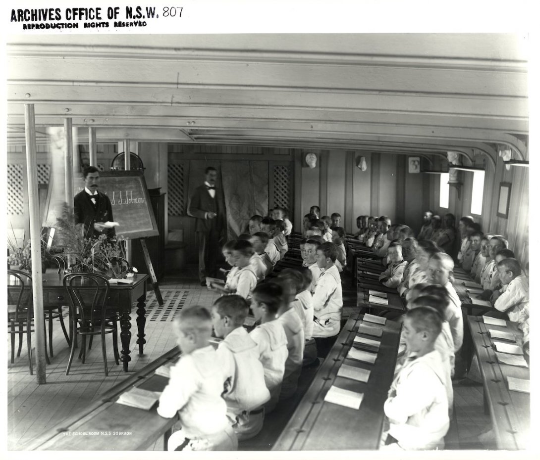 Classroom on a ship with many young boys sitting at long curved desks and two teachers standing at the front