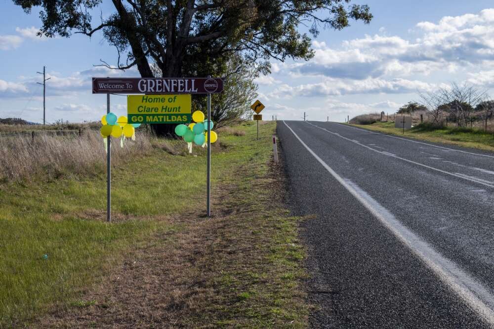 A country road with a road sign to the left that reads 'Welcome to Grenfell. Home of Clare Hunt. Go Matildas.'