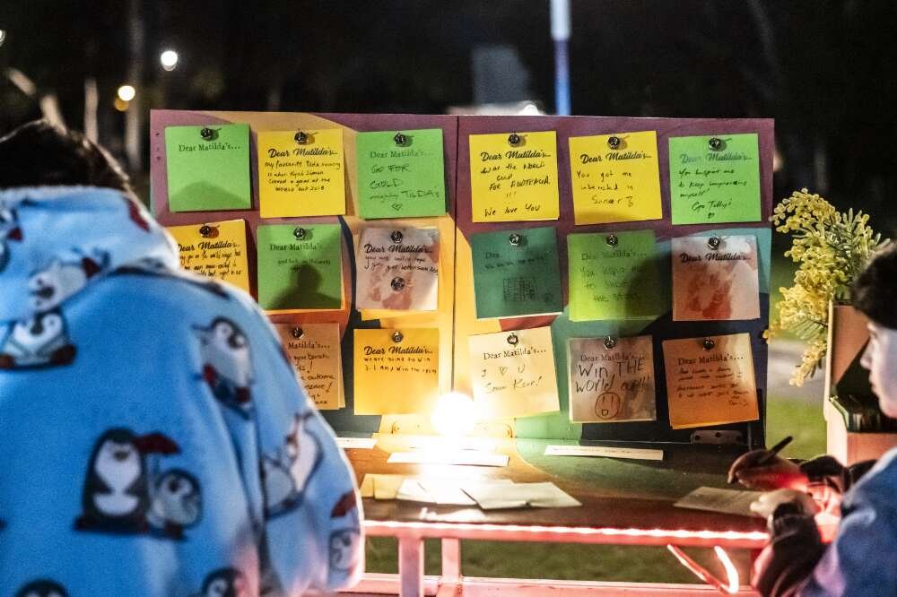 The backs of two people standing infront of a lit up corkboard that shows messages of support for the Matildas.
