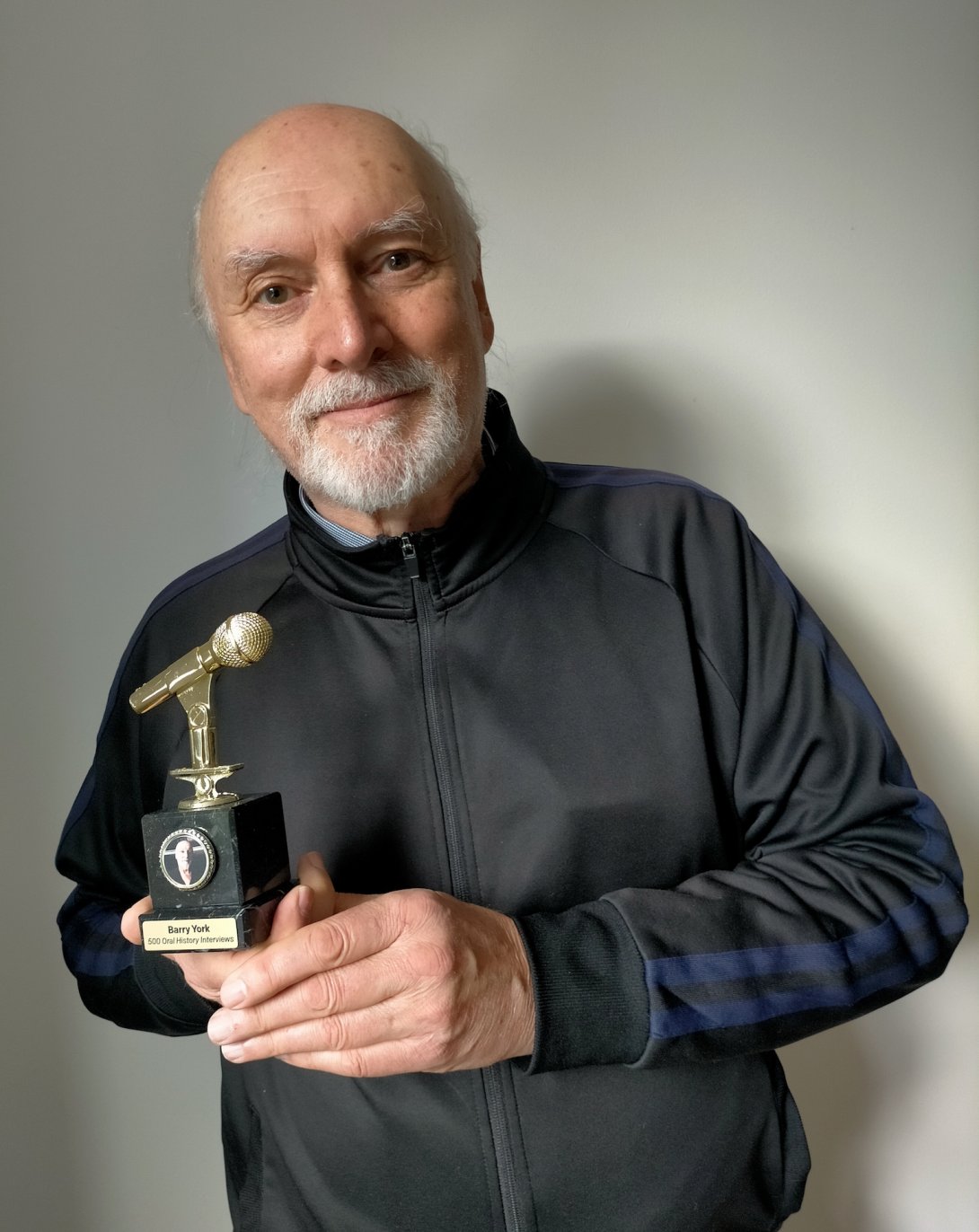 Middle-aged man with a white beard smiling and holding up a trophy with a golden microphone that reads 'Barry York 500 Oral History Interviews'