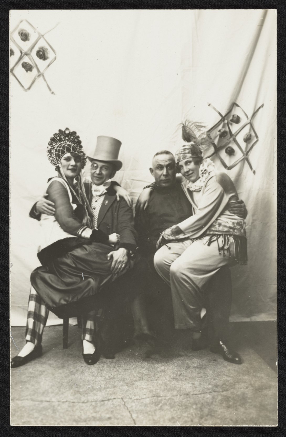 Photo of two women sitting on the laps of two men, all four wearing elaborate clothes with various headdresses