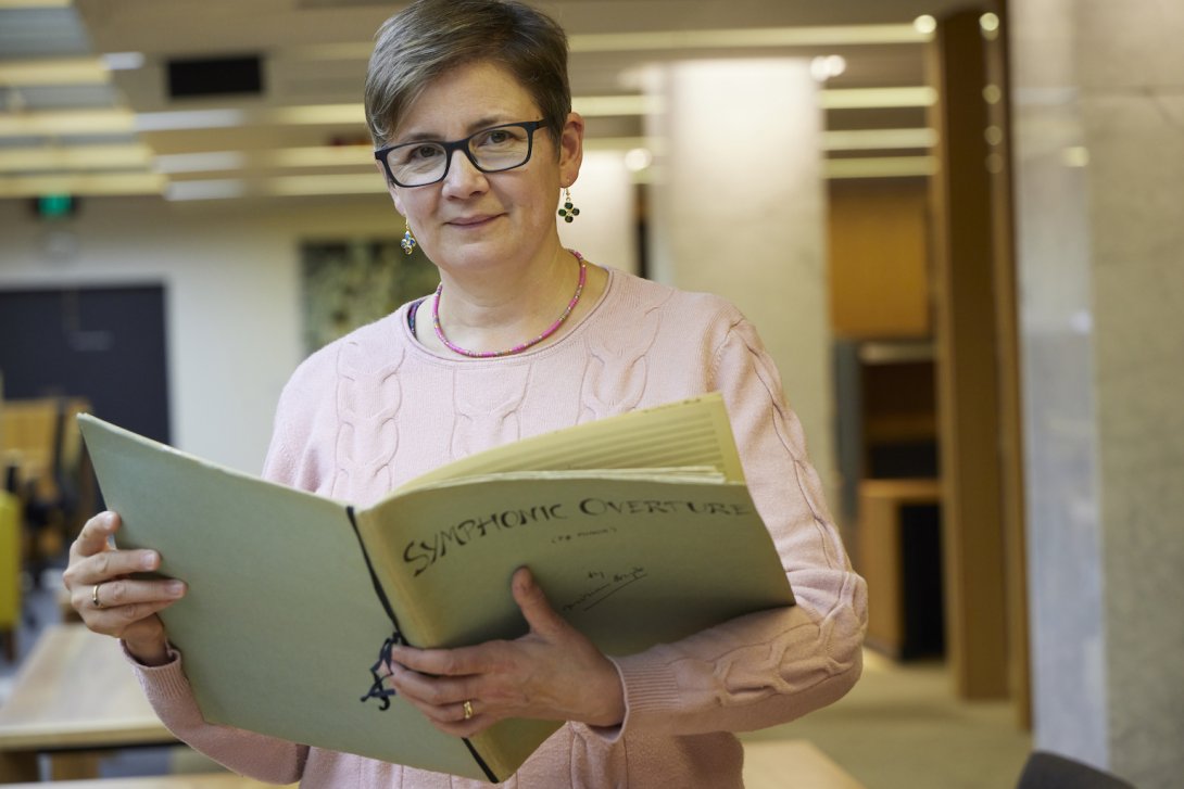 Celia Craig, a woman with very short brown hair, smiling and holding large book of sheet music with the title 'Symphonic Overture'