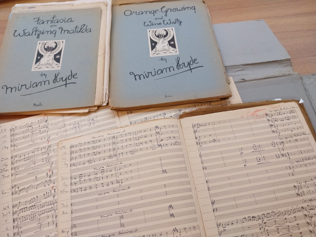 Sheet music books, some open and some closed, spread over a wooden table. The closed books' covers read 'Fantasia on Waltzing Matilda by Miriam Hyde' and 'Orange Growing and Wine Waltz by Miriam Hyde'