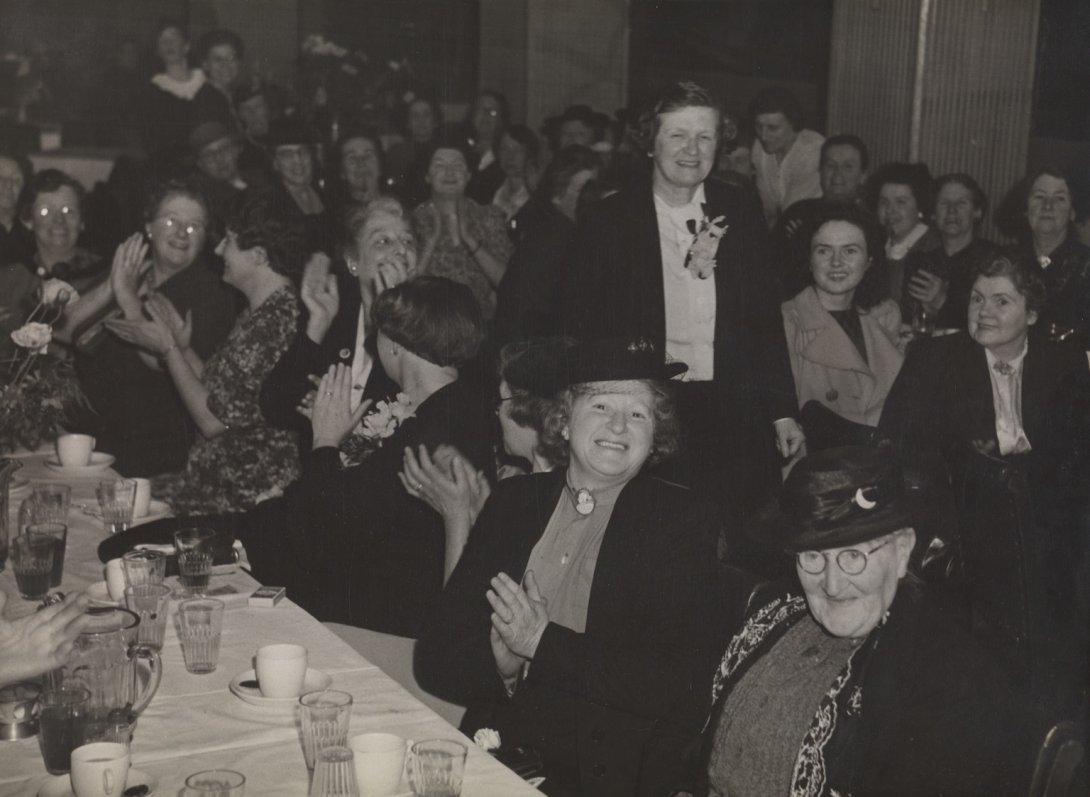 Jessie Street walking between tables with dozens of women seated and clapping for her