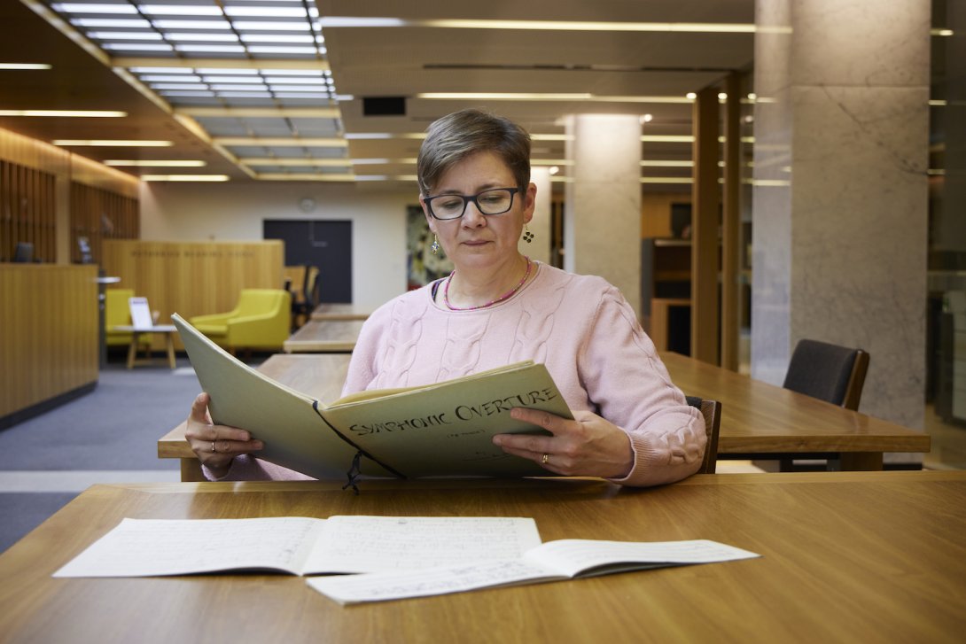 Celia Craig, a woman with very short brown hair, reading a large book of sheet music with the title 'Symphonic Overture' at a table in the Library's Special Collections Reading Room